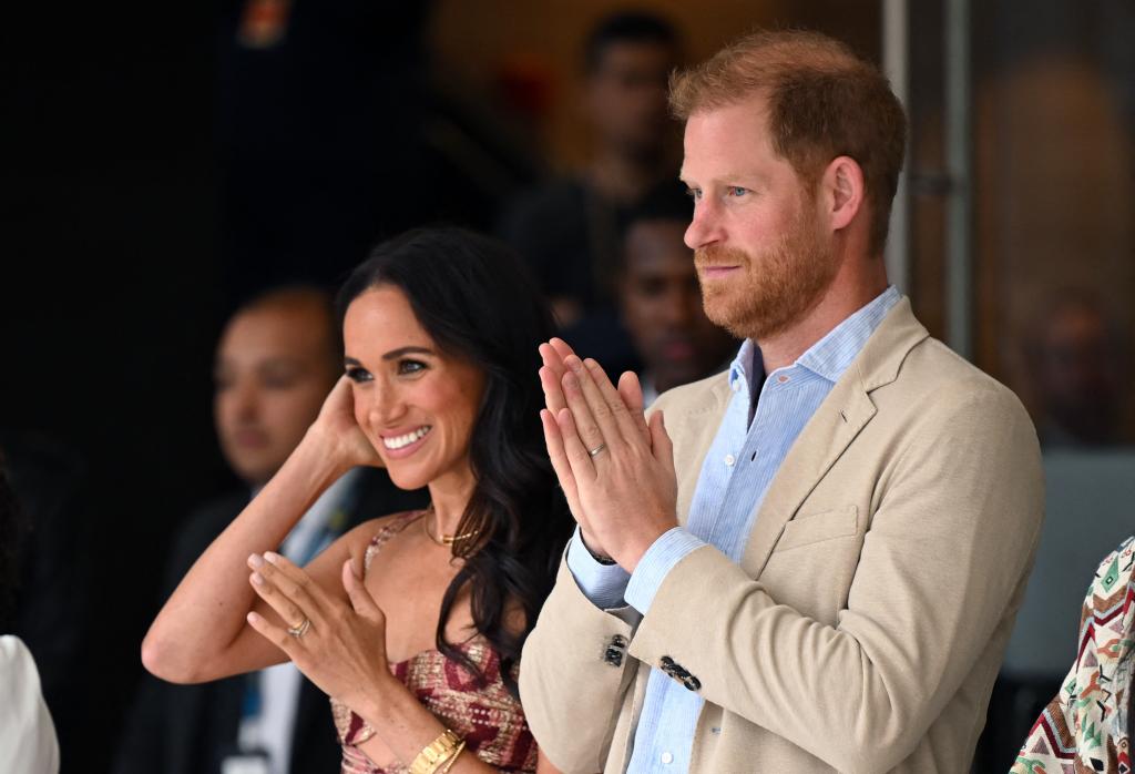 Meghan Markle, in a patterned summer dress, stands next to Prince Harry, in a cream blazer and blue shirt.