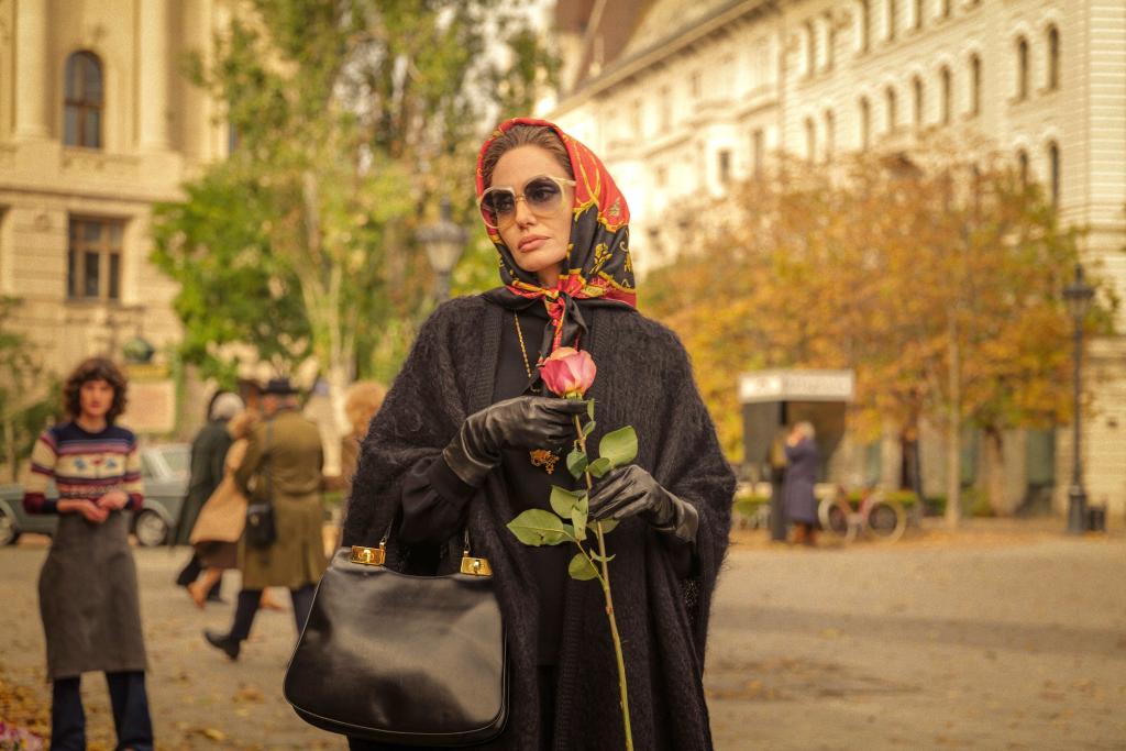 Angelia Jolie in a red headscarf, sunglasses, and a black coat, carrying a rose.